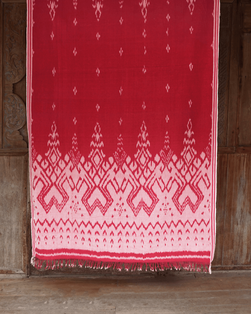 Ręcznie wytwarzana tkanina ikat z Bali, w odcieniach czerwieni i bieli, zawieszona na tle drewnianego domku na bali. Tkanina ozdobiona tradycyjnymi geometrycznymi i wzorami, podkreślającymi unikalność rękodzieła.