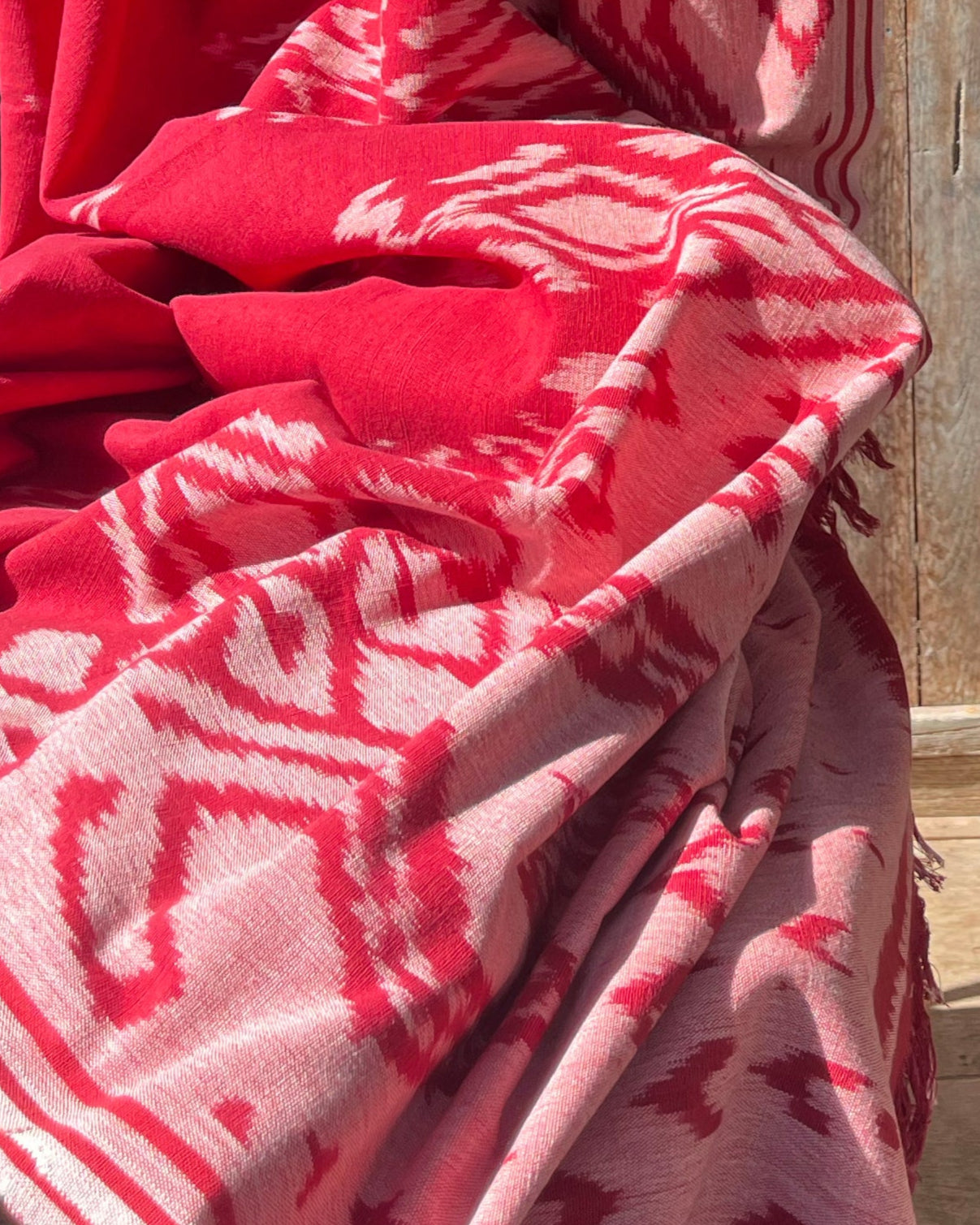 Ręcznie wytwarzana tkanina ikat z Bali, w odcieniach czerwieni i bieli, położona na tle drewnianego domku na bali. Tkanina ozdobiona tradycyjnymi geometrycznymi i wzorami, podkreślającymi unikalność rękodzieła.