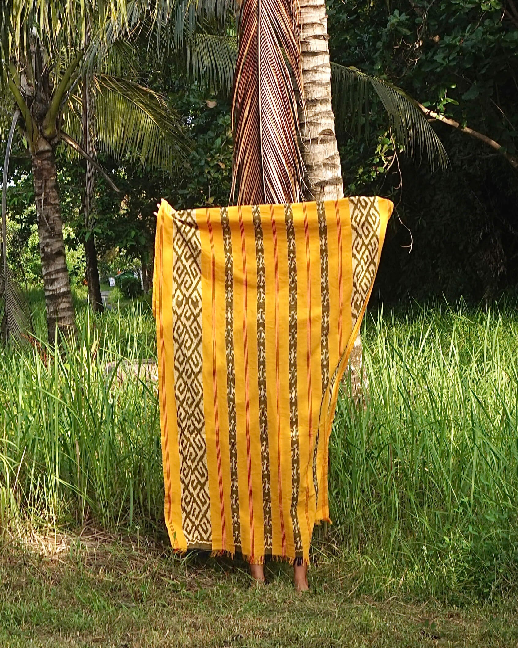 Ręcznie wytwarzana tkanina Balileo ikat z Bali, w odcieniach żółci, czerni i bieli, zawieszona na tle tropikalnych roślin. Tkanina ozdobiona tradycyjnymi geometrycznymi i wzorami, podkreślającymi unikalność rękodzieła.