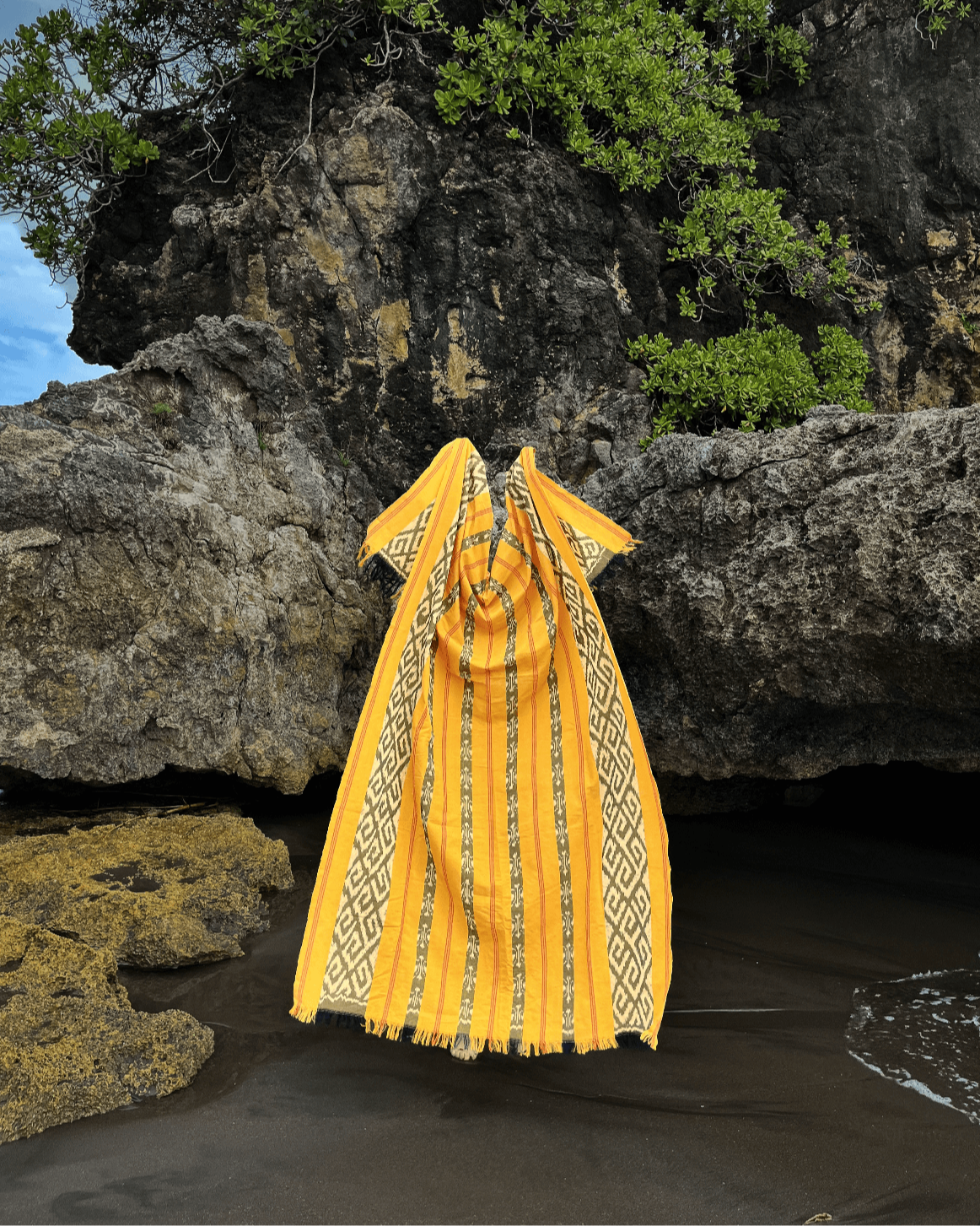 Ręcznie wytwarzana tkanina Balileo ikat w odcieniach żółci, czerni i bieli, zawieszona na skalistej plaży na Bali. Tkanina ozdobiona tradycyjnymi geometrycznymi i wzorami, podkreślającymi unikalność rękodzieła.