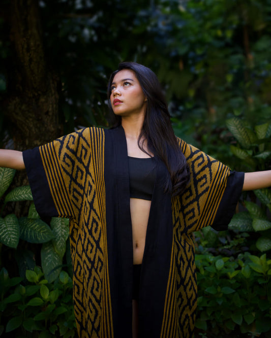 Kobieta kimonie z ręcznie wytwarzanej tkaniny Balileo ikat, w odcieniach żółci, z charakterystycznymi czarnymi, geometrycznymi wzorami, stojąca na tle tropikalnych roślin na Bali. Tkanina zdobiona tradycyjnymi wzorami podkreśla unikalność rękodzieła.