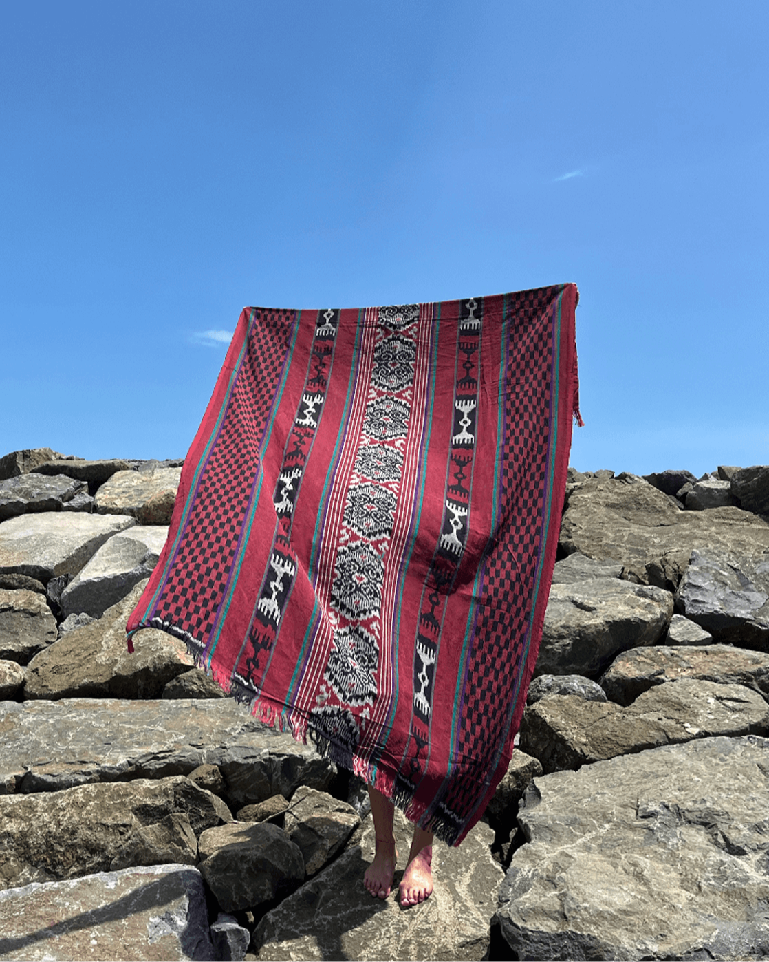 Ręcznie wytwarzana tkanina ikat z Bali, w odcieniach czerwieni, czerni i bieli, zawieszona na tle kamienistego brzegu i błękitnego nieba. Tkanina ozdobiona tradycyjnymi geometrycznymi i wzorami, podkreślającymi unikalność rękodzieła.