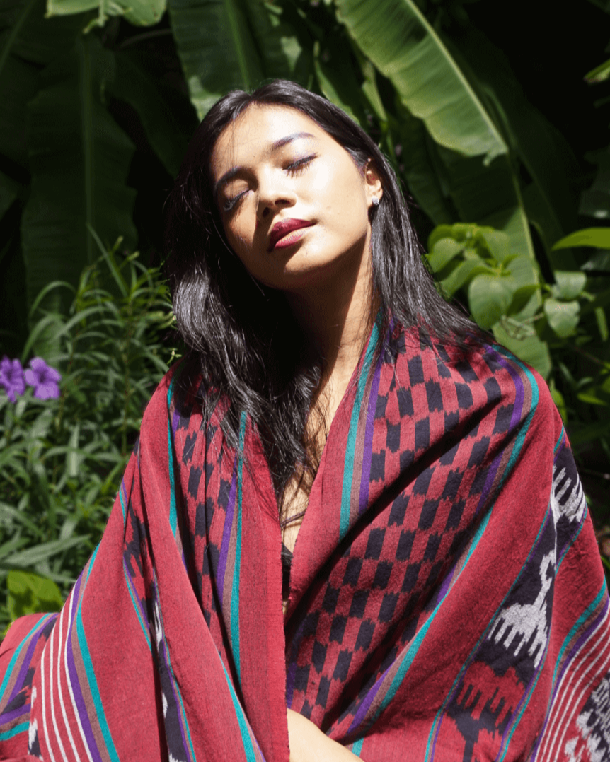 Kobieta owinięta ręcznie wytwarzaną tkaniną ikat z Bali, w odcieniach czerwieni, czerni i fioletu, siedząca w tropikalnym otoczeniu z dużymi liśćmi bananowca w tle. Tkanina zdobiona tradycyjnymi wzorami podkreśla unikalność rękodzieła.