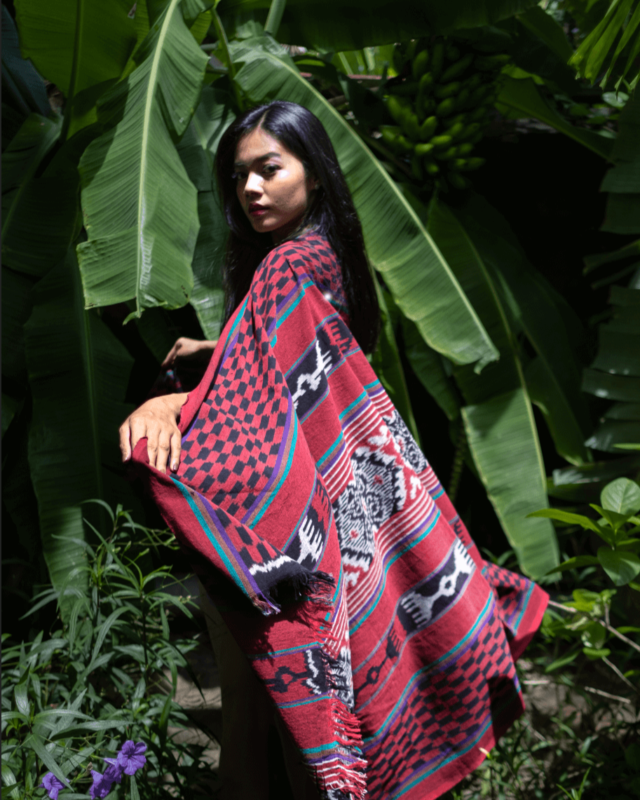 Kobieta owinięta ręcznie wytwarzaną tkaniną ikat z Bali, w odcieniach czerwieni, czerni i fioletu, stojaca w tropikalnym otoczeniu z dużymi liśćmi bananowca w tle. Tkanina zdobiona tradycyjnymi wzorami podkreśla unikalność rękodzieła.