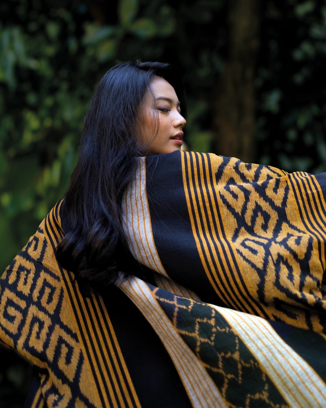 Kobieta stojąca tyłem kimonie z ręcznie wytwarzanej tkaniny Balileo ikat, w odcieniach żółci, z charakterystycznymi czarnymi, geometrycznymi wzorami, stojąca na tle tropikalnych roślin na Bali. Tkanina zdobiona tradycyjnymi wzorami podkreśla unikalność rękodzieła.